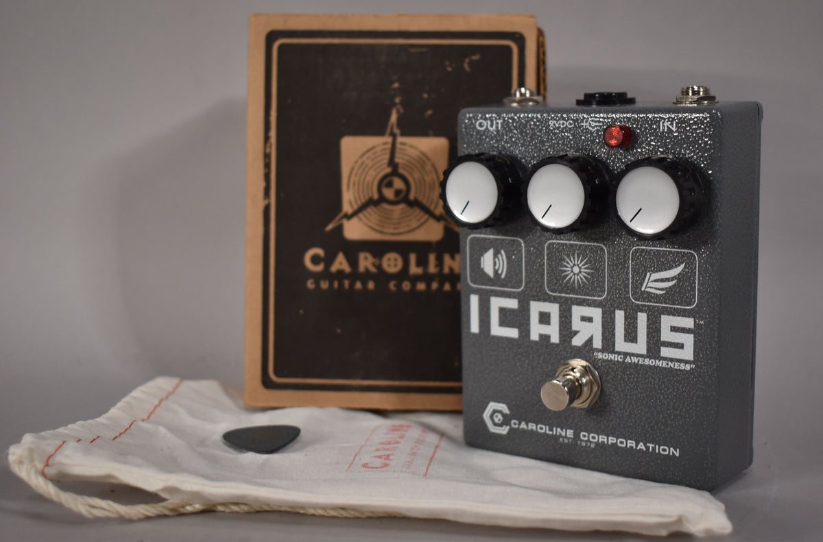 A dark gray textured pedal with three white-capped black knobs, a red LED, and a silver footswitch, shown with its brown box.