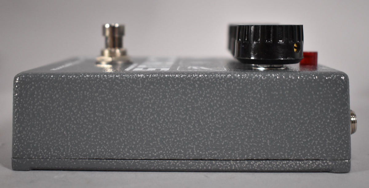 The gray speckled pedal is viewed from the side, showing a silver footswitch, a black knob, and a red indicator light.