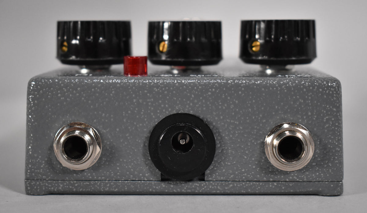 The gray speckled pedal is viewed from the back, showing three black knobs, a red button, two input jacks, and a power input.