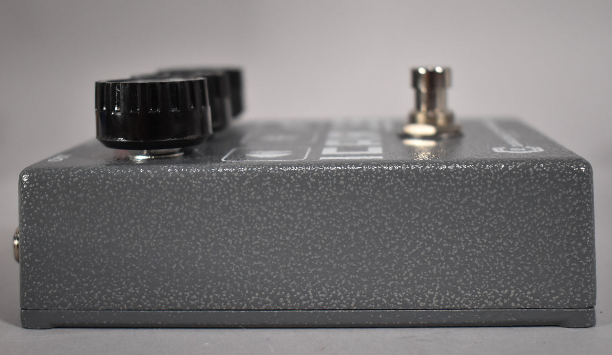 The gray, speckled effect pedal is viewed from the side, showing three black knobs and a silver input jack on the left.