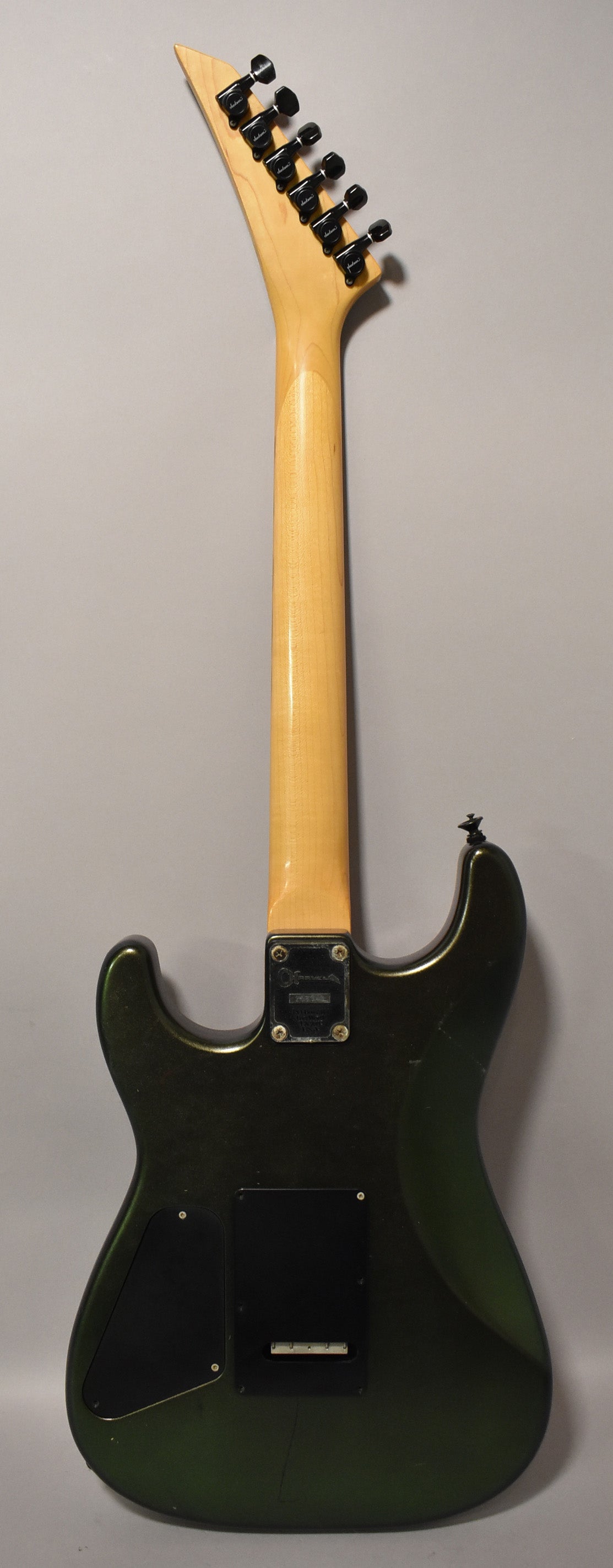 The back of a dark green metallic electric guitar, showing the maple neck, black tuners, and control cavity covers.