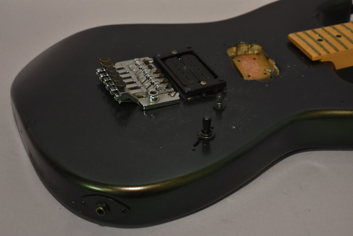 Close-up of a dark metallic green guitar body with a chrome tremolo, black pickup ring, empty pickup cavity, and two loose control knobs.