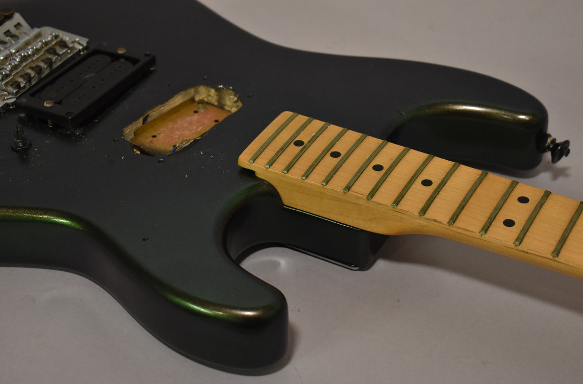 The dark green metallic guitar body is missing a neck pickup, with a maple neck and fretboard resting beside it.