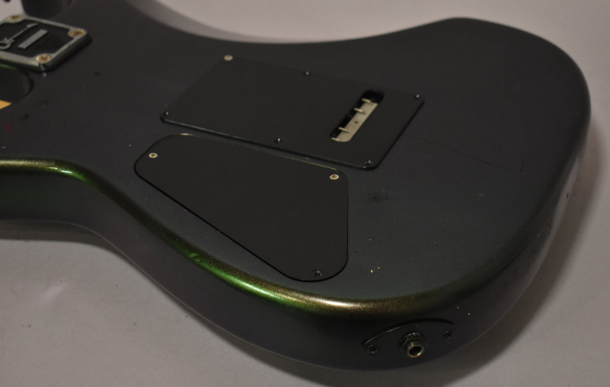 The guitar's dark gray body with green metallic flake is seen from the back, showing two control cavity covers and the output jack.