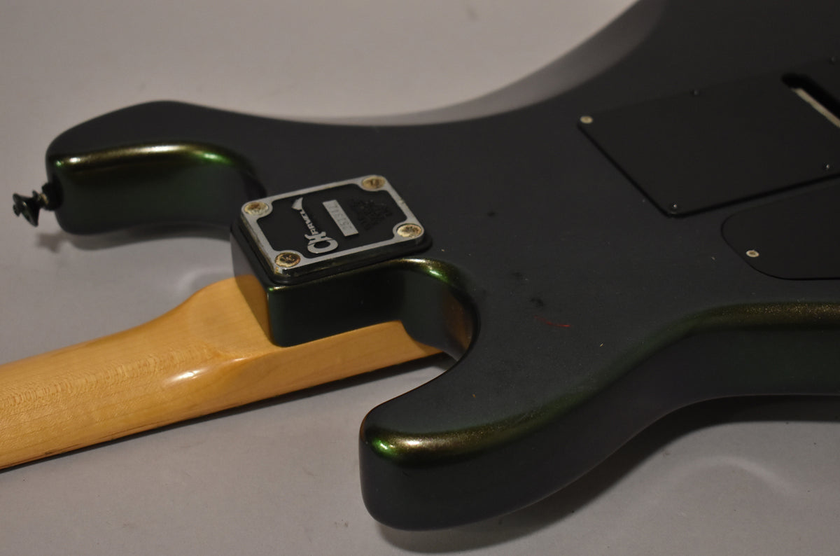 The back of a dark green electric guitar body with a maple neck, showing a metal neck plate and black control cavity covers.
