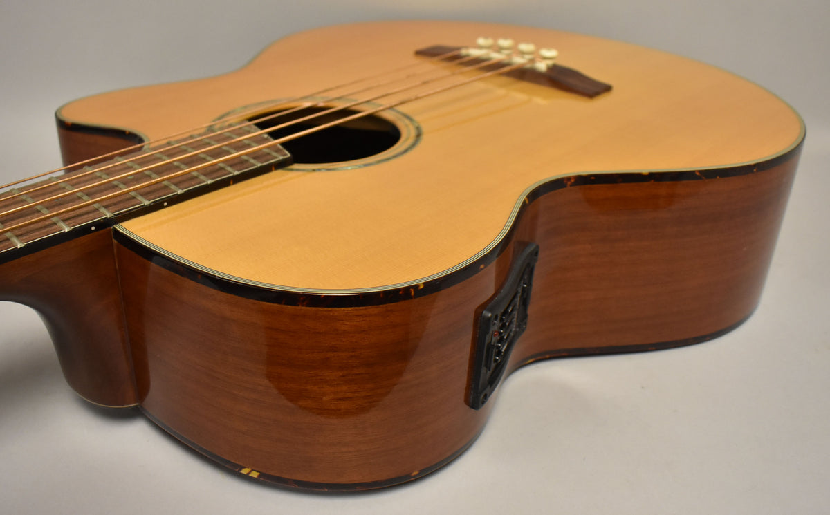 Angled view of the natural finish acoustic bass, showing the light wood top, dark wood sides, soundhole, bridge, and a black preamp control panel.