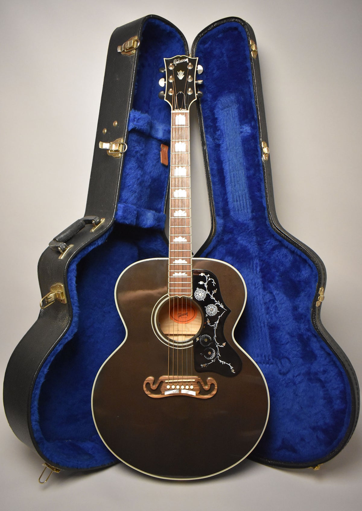 A full-length view of a glossy trans black jumbo acoustic guitar with a decorative pickguard and a blue-lined hard case.