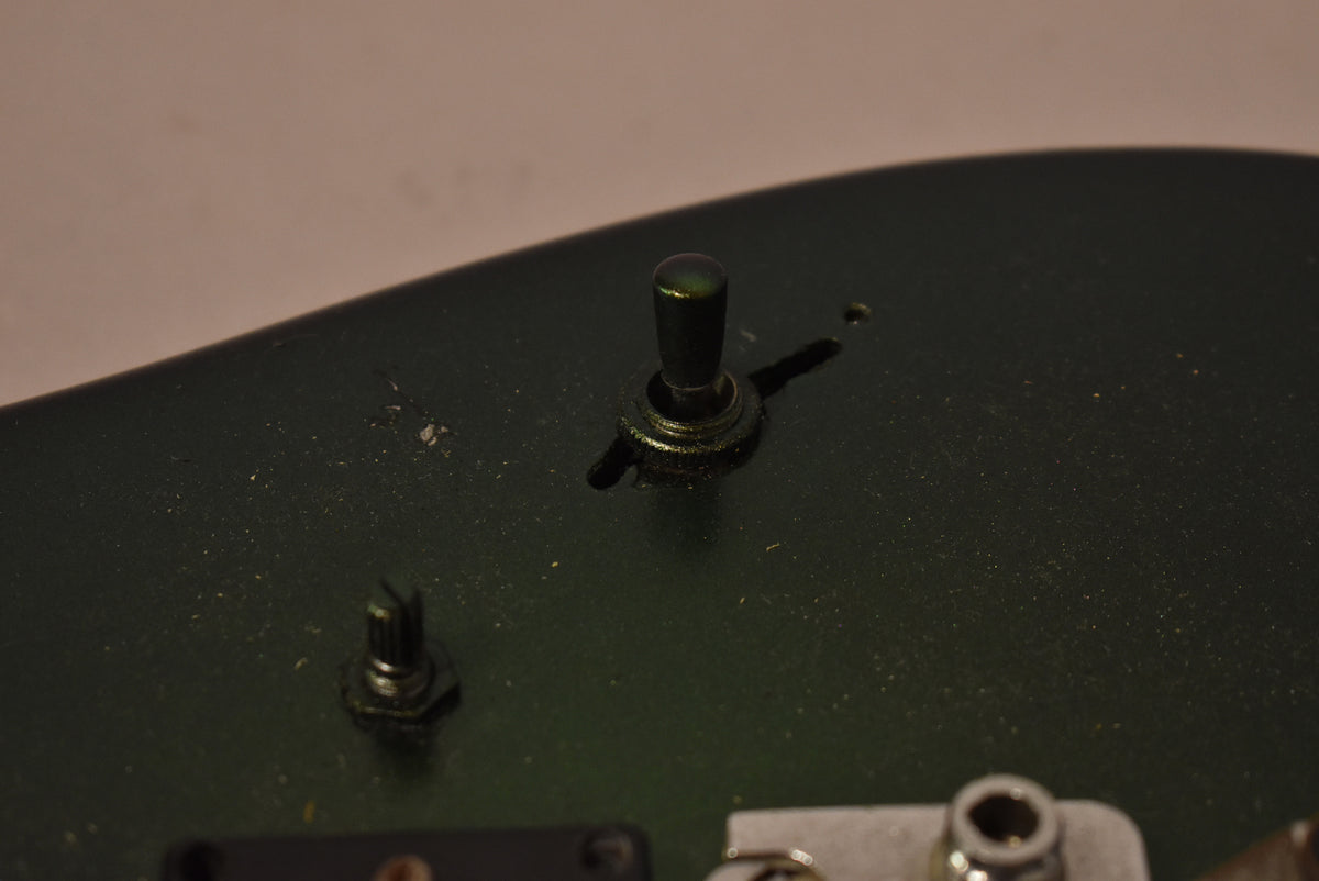 Close-up of the dark green guitar body, showing a black toggle switch, a stripped pot shaft, and a jack socket.
