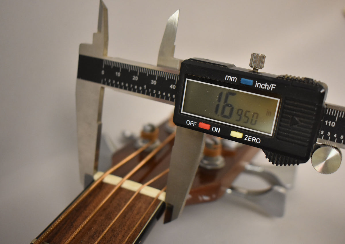 A digital caliper measures the nut width of the natural finish acoustic bass, showing 1.6950 inches.