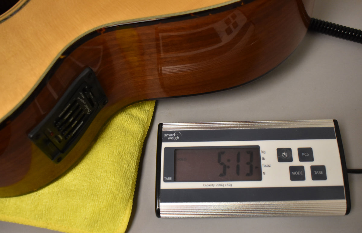 The natural finish acoustic bass, with a black preamp visible on its side, rests on a yellow cloth next to a digital scale displaying "5:13".