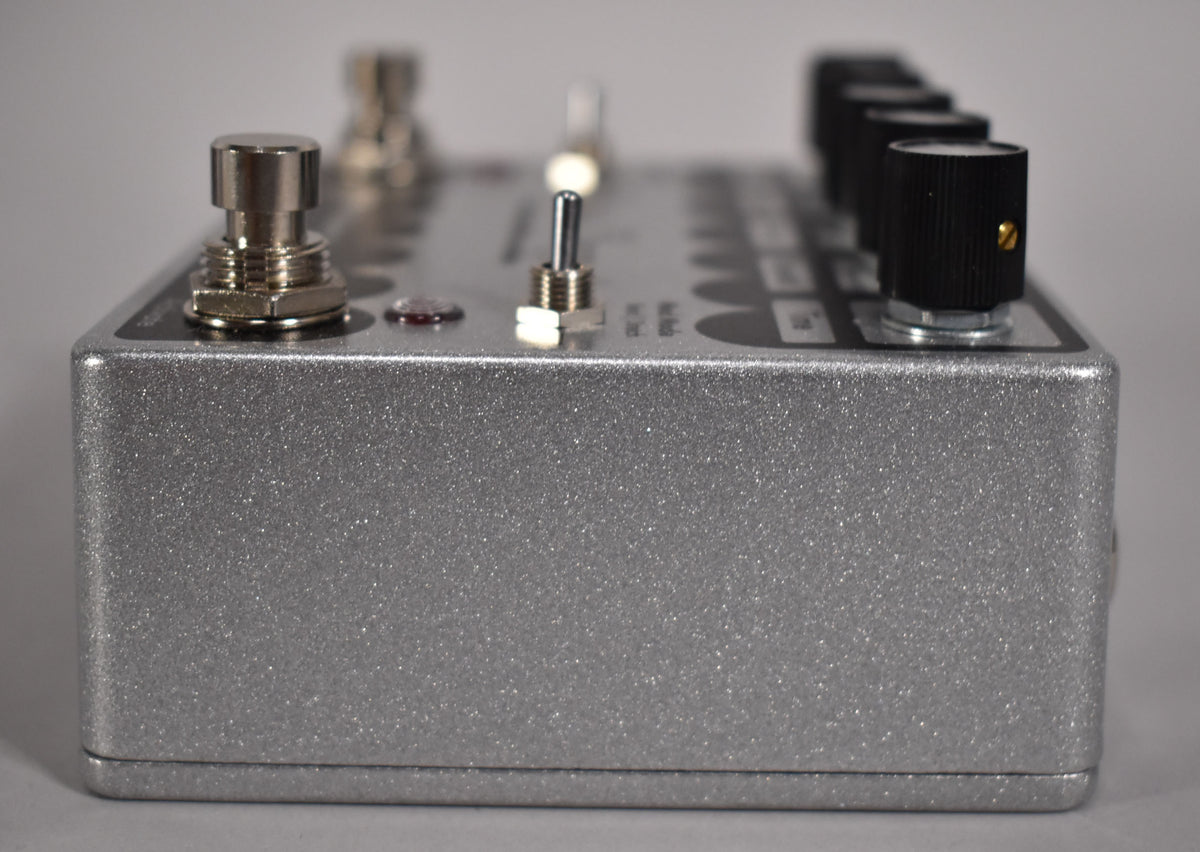 A close-up, angled view of a silver glitter-finished guitar pedal with a footswitch, toggle switch, and several black control knobs.