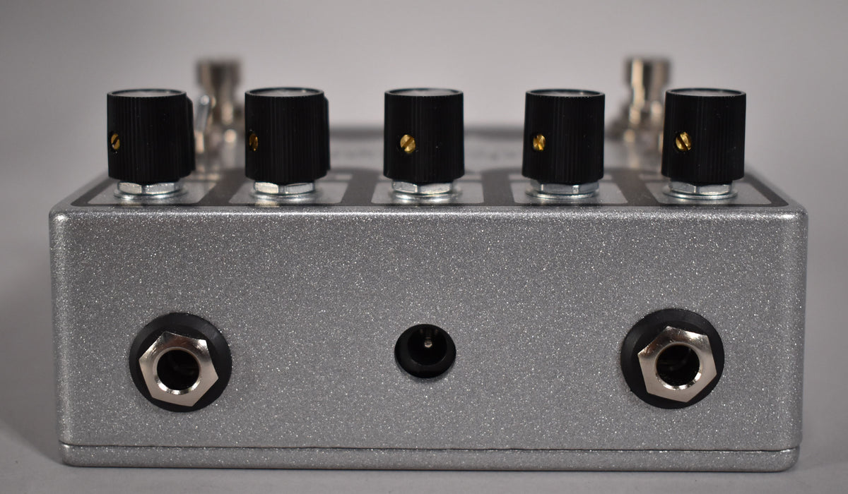 The silver glitter-finish pedal is viewed from the back, showing two input jacks, a power input, and five black control knobs.