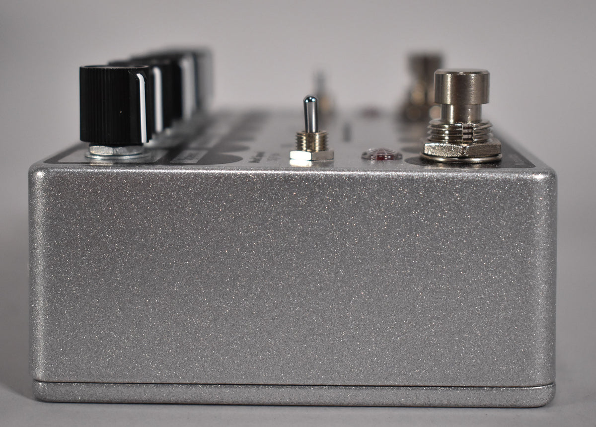 The silver glitter-finish pedal is viewed from the right side, showing black knobs, a metal toggle switch, and a footswitch.