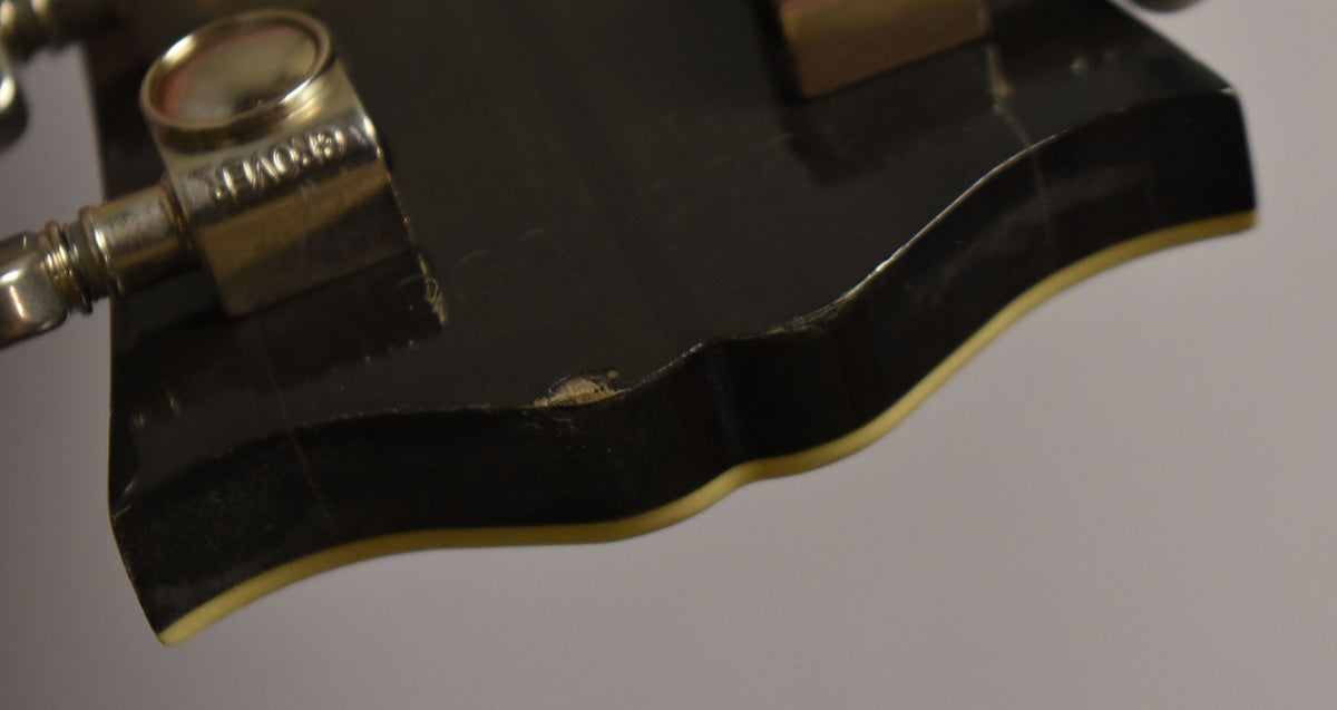 Close-up of the black headstock's edge, showing a small chip in the finish and binding, with a chrome tuning peg visible.