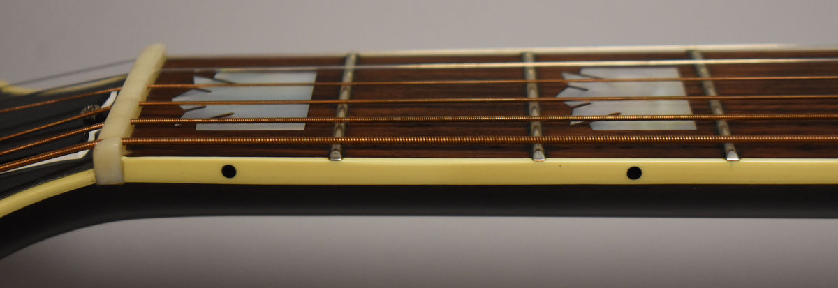 Close-up, side view of the guitar's neck, showing the nut, first few frets, and trapezoid mother-of-pearl inlays on the dark fretboard.