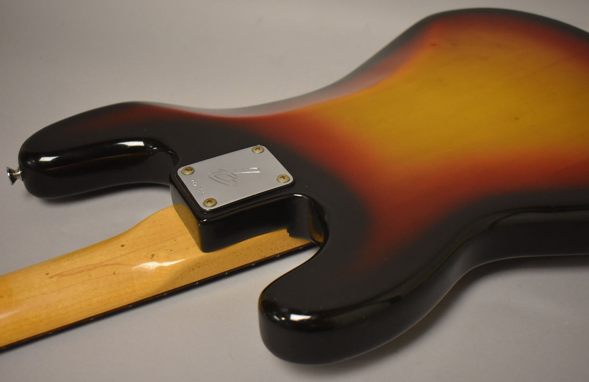 The bass's sunburst body and neck joint are visible from the back, showing a chrome neck plate and wear on the maple neck.