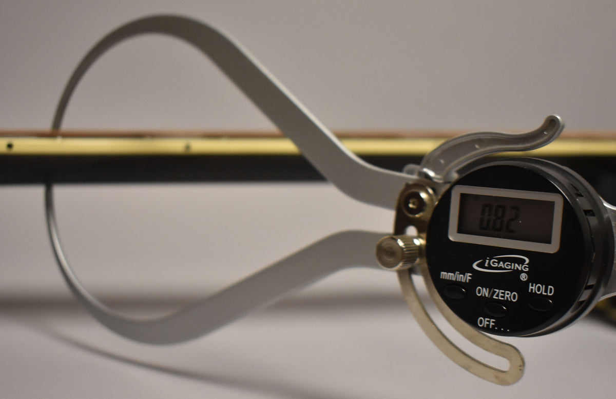 Close-up of the guitar's black neck and light brown fretboard, with a digital caliper measuring the action at 0.082 inches.