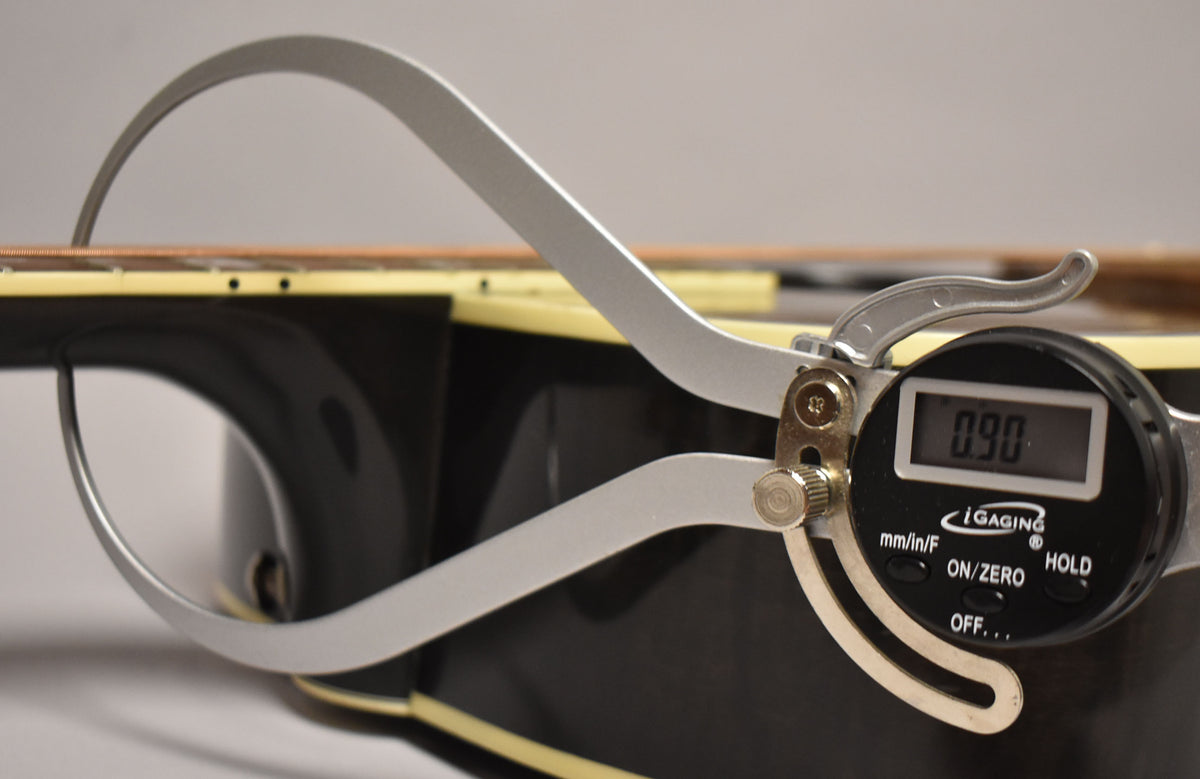 A digital caliper measures the guitar's nut width at 0.90, with the black gloss finish and cream binding visible.