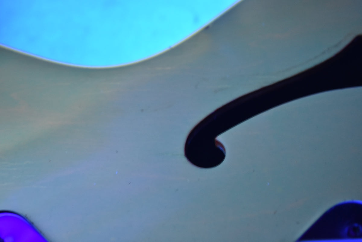 Close-up of a guitar's f-hole, showing the light-colored body and dark f-hole against a bright blue background.