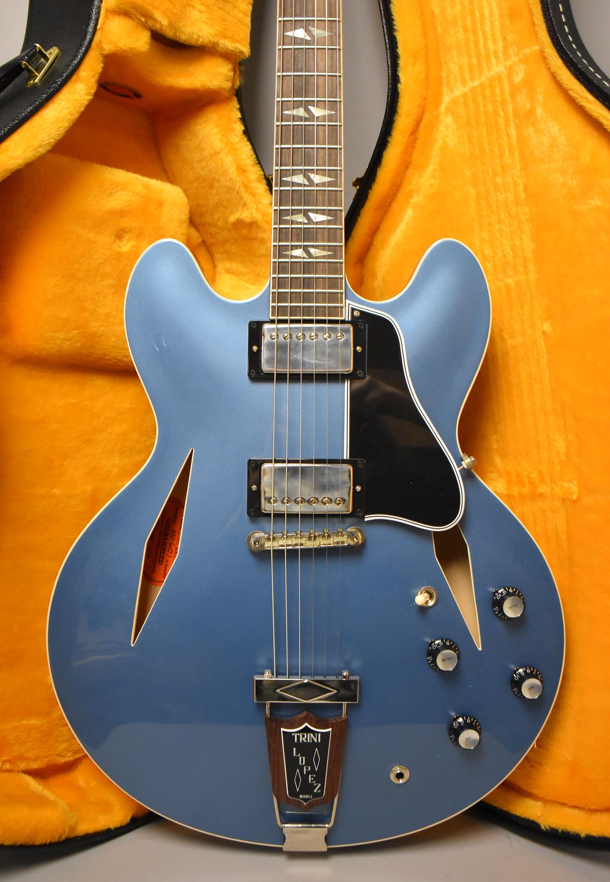 A Pelham Blue semi-hollowbody electric guitar with f-holes, two humbuckers, and a Trini Lopez tailpiece, shown in its open orange-lined case.