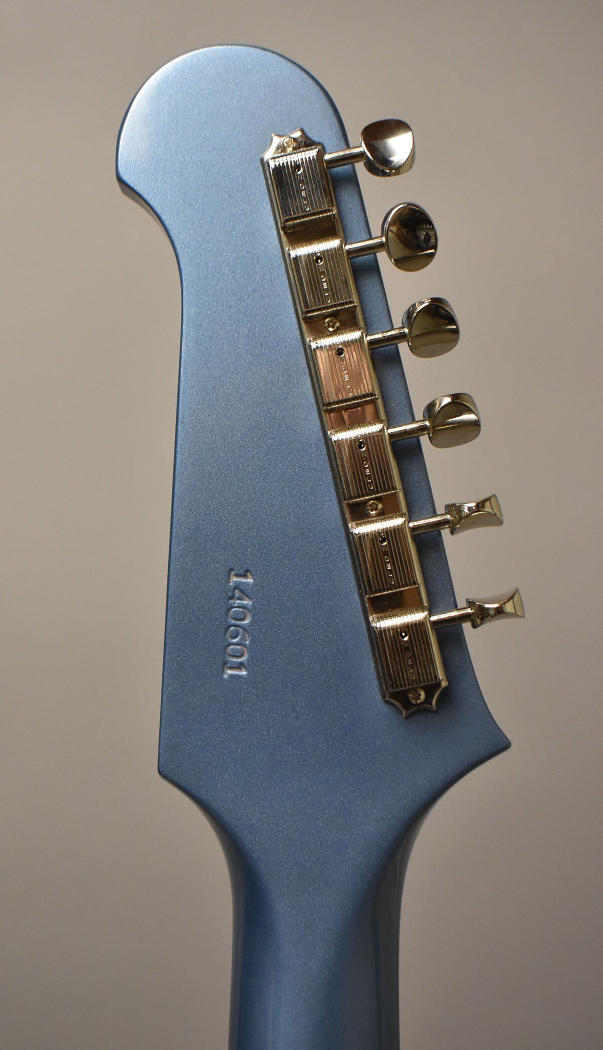The back of a Pelham Blue headstock with a matte finish, showing six gold-plated tuners and an embossed serial number.