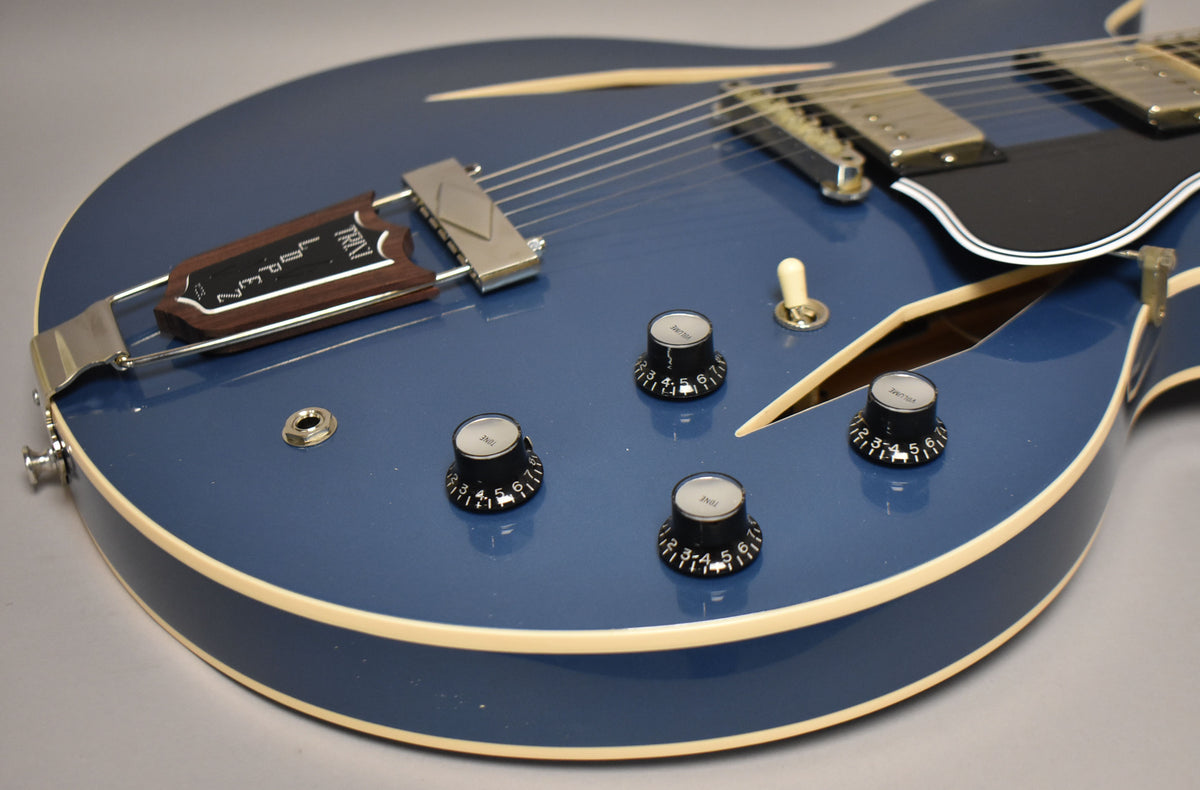 The Pelham Blue guitar body, with cream binding, shows four black control knobs, a chrome trapeze tailpiece, and an f-hole.