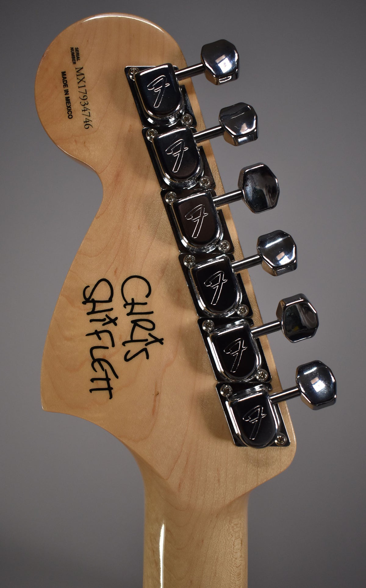 Rear view of a natural wood headstock with six chrome tuners, angled slightly left, showing "Chris Shiflett" in black script.