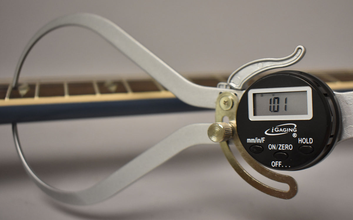 A digital caliper measures the Pelham Blue guitar's neck relief at 0.01 inches, showing the fretboard and frets.