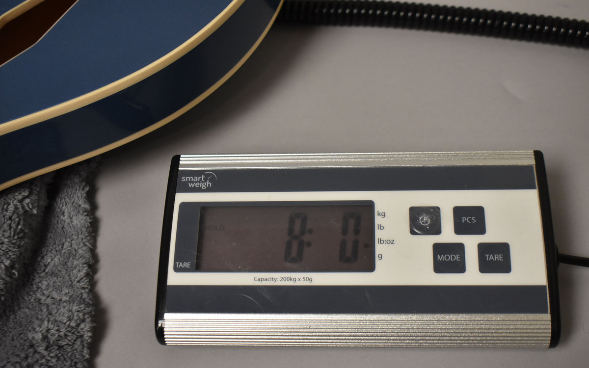 The Pelham Blue guitar body's lower bout and binding are visible, resting on a grey cloth next to a digital scale reading "0.0".