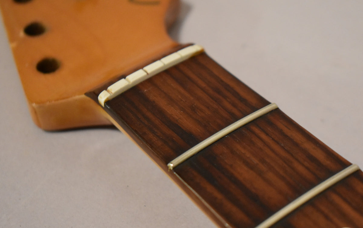 Close-up of the dark rosewood fretboard and light frets, with a bone nut and three tuner holes visible on the light brown headstock.