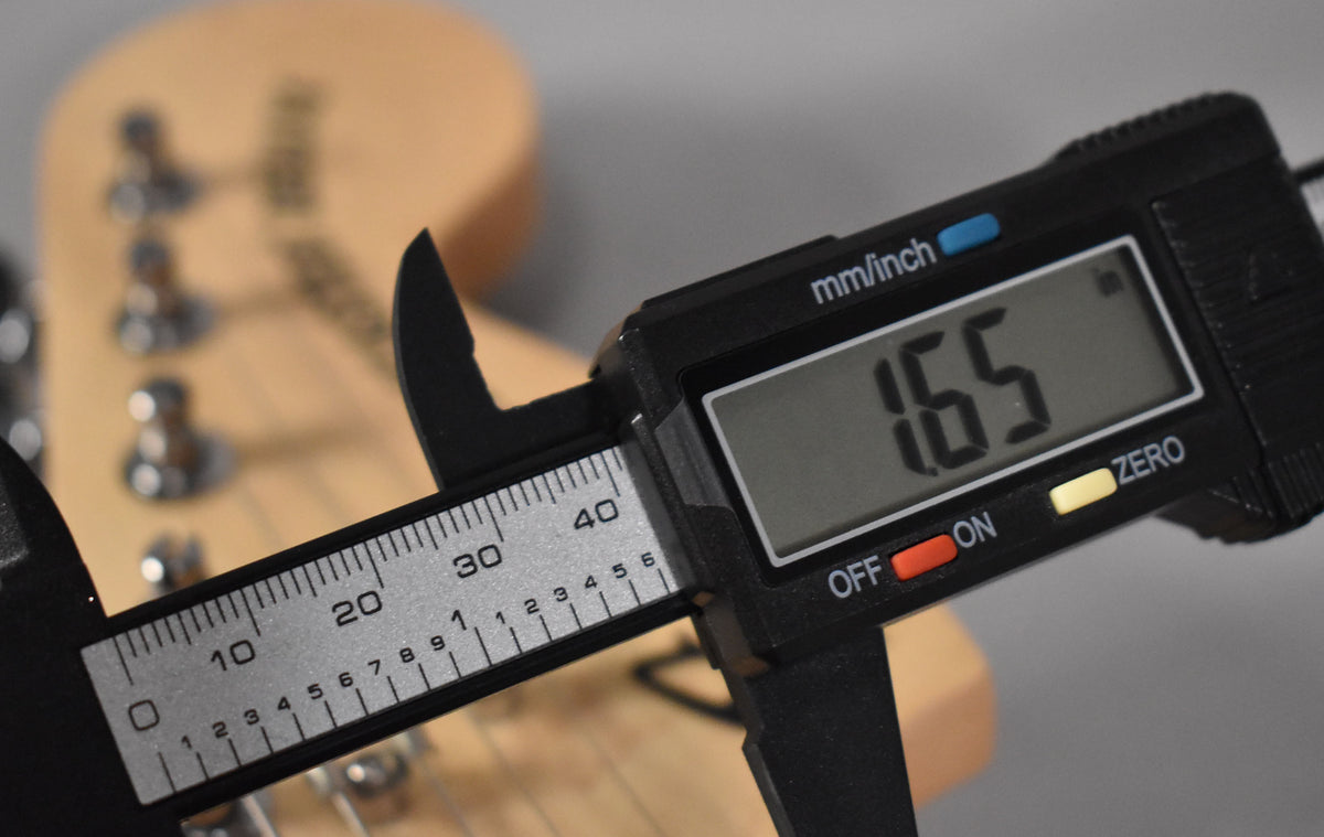 A digital caliper measures the guitar's nut width at 1.65 inches, with the headstock and tuning pegs blurred in the background.