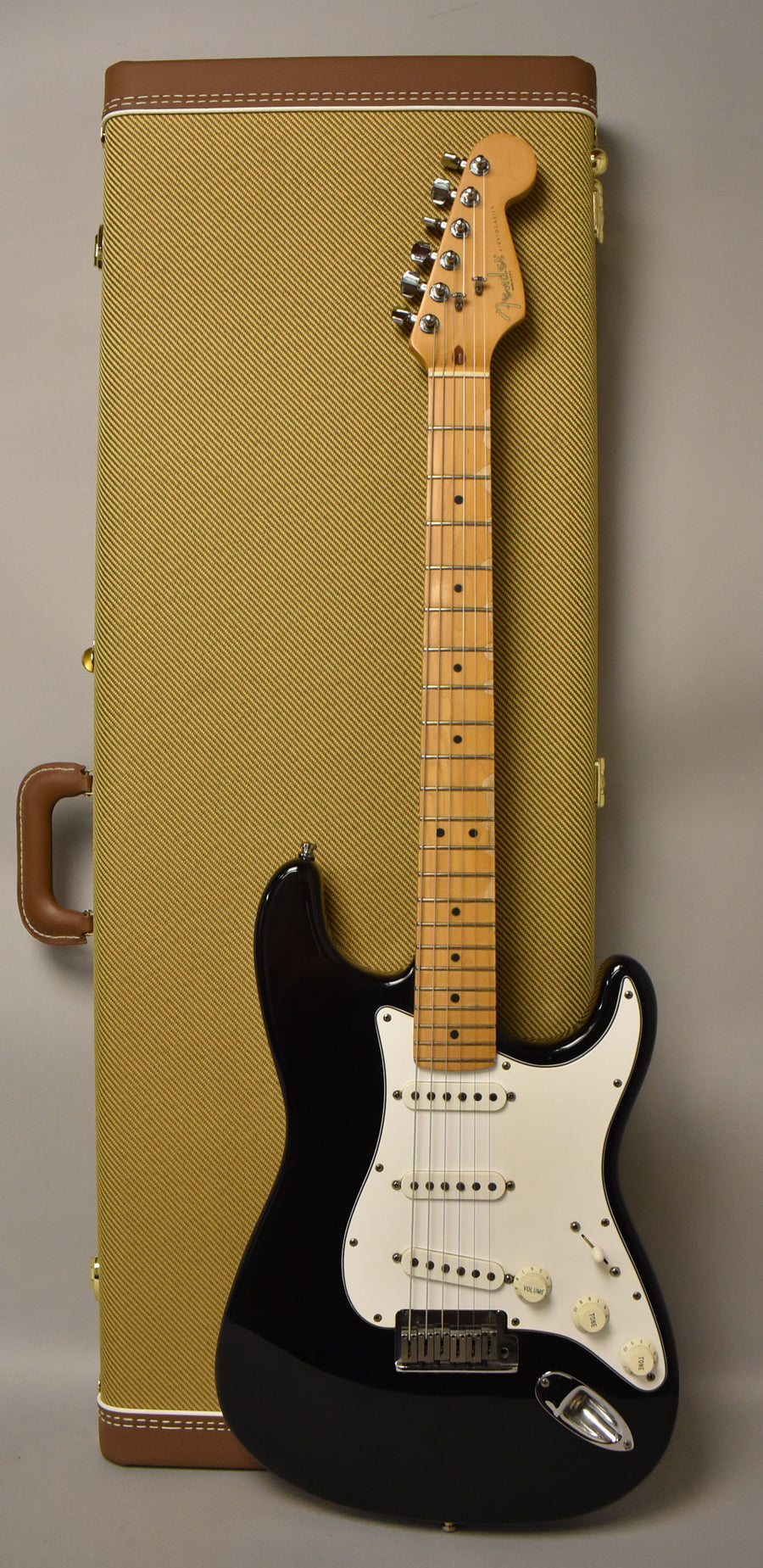 A black Stratocaster with a white pickguard and maple neck stands upright against a tweed hard shell case.