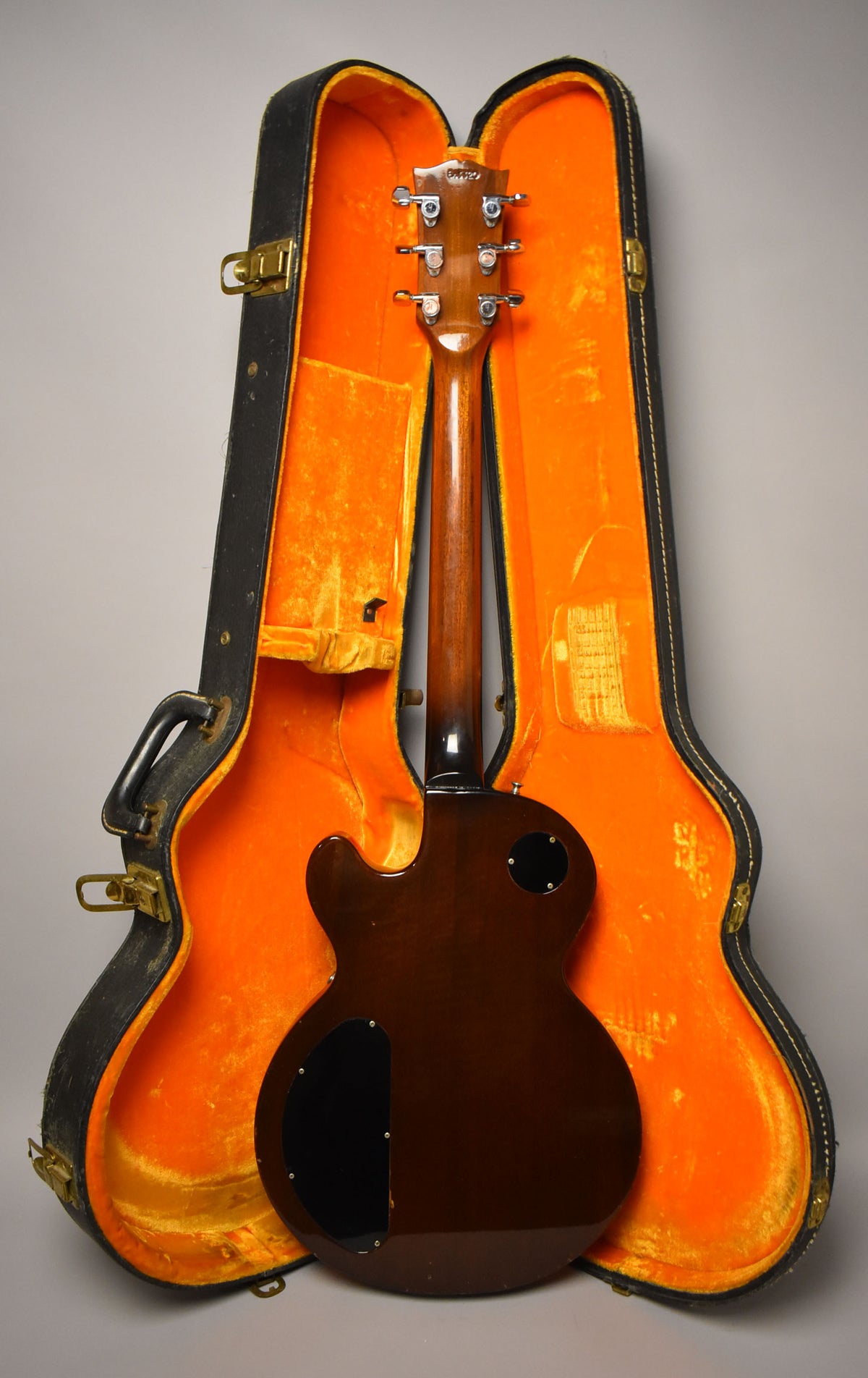 The guitar's dark walnut back and neck are visible, with a black control cavity cover and a round access plate, resting in an open orange-lined case.