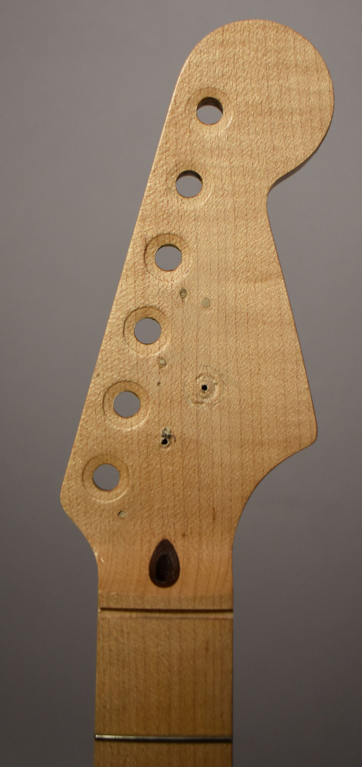 Close-up of a light maple Stratocaster headstock with five tuner holes, a dark tear-drop string tree, and visible wood grain.