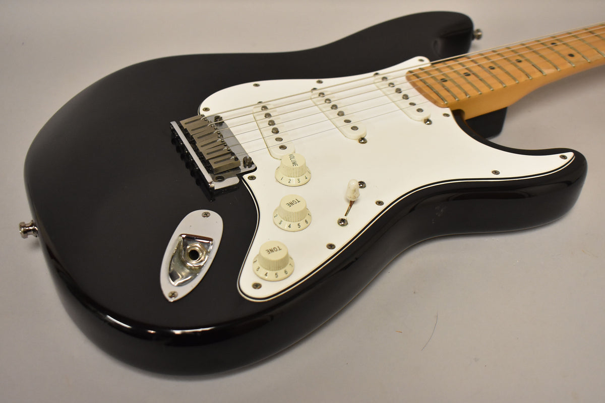 Black electric guitar, angled from the lower bout, showing the white pickguard, three single-coil pickups, and three cream control knobs.