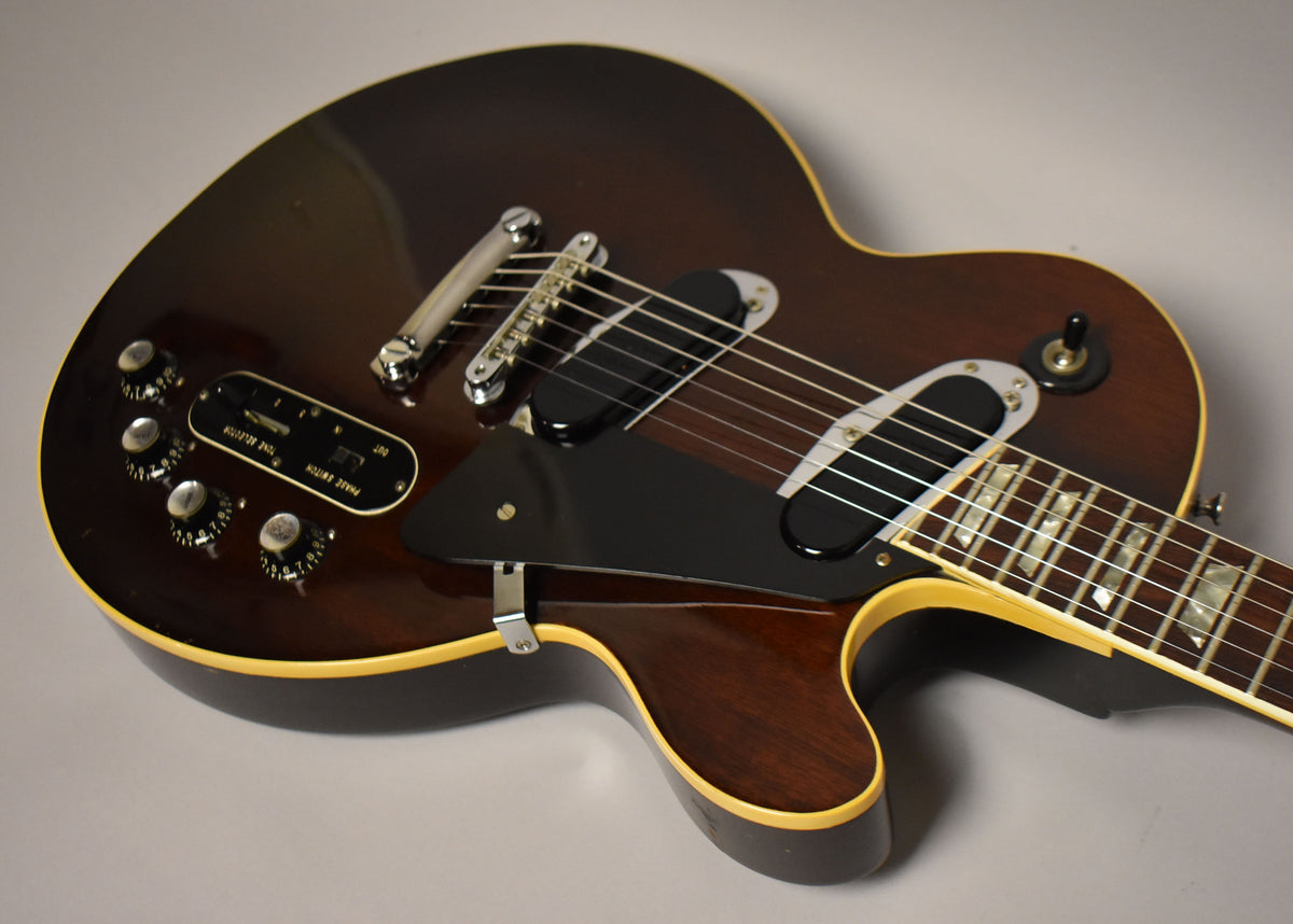The dark walnut guitar body, with cream binding, shows a black pickguard, two humbuckers, and a control plate with four knobs and two switches.