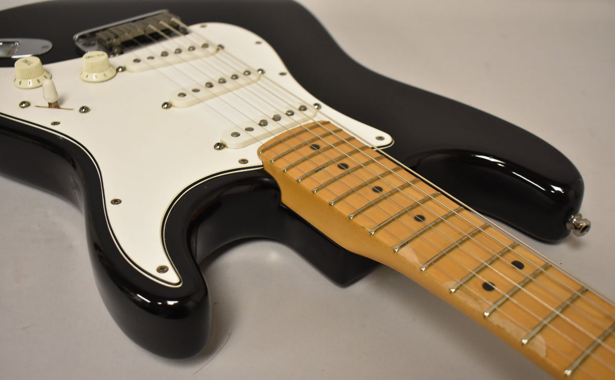 The black electric guitar, with a white pickguard and maple neck, is angled to show the body and lower fretboard.