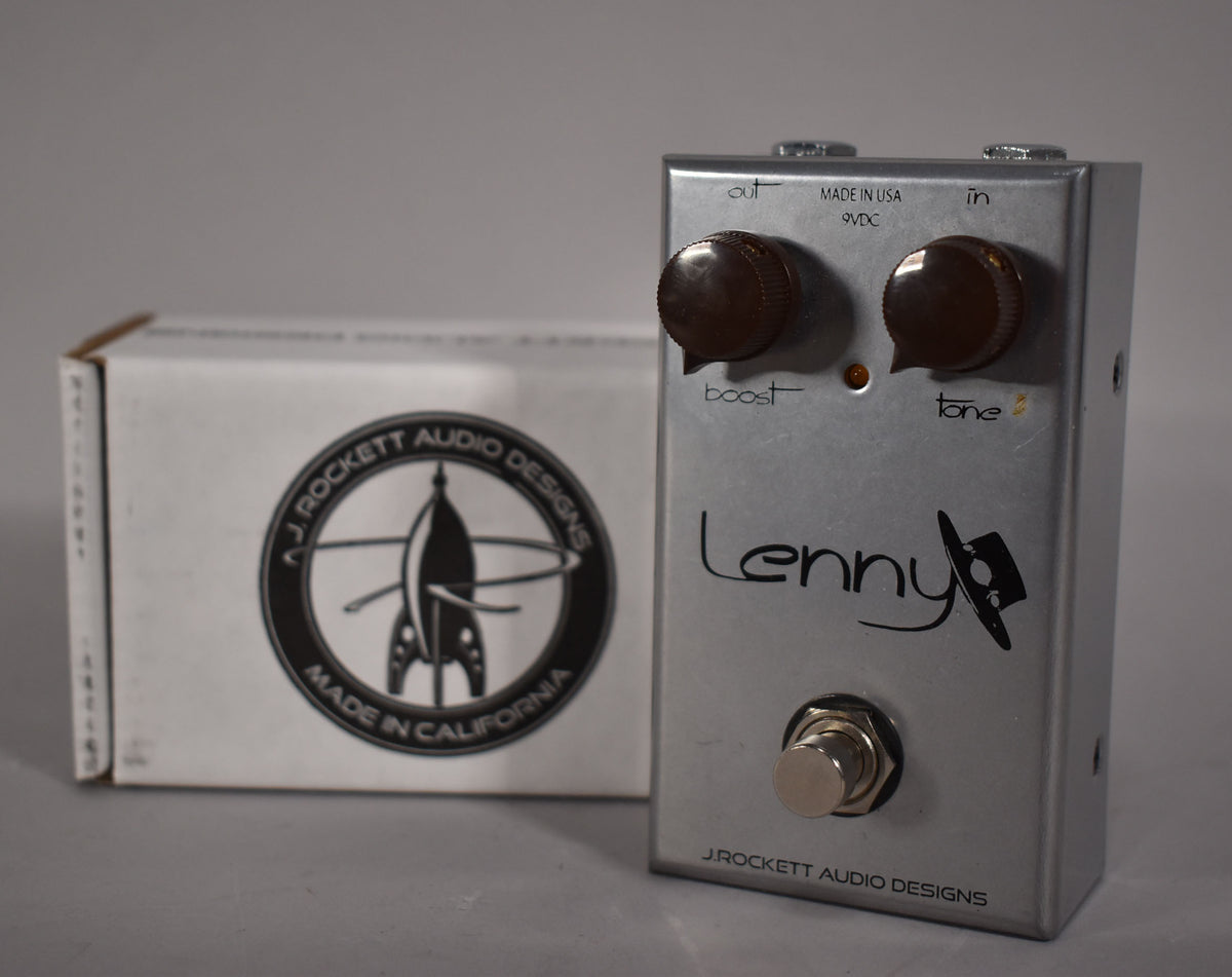 A silver effects pedal with two brown knobs, a footswitch, and a white box with a rocket logo behind it, viewed from the front.