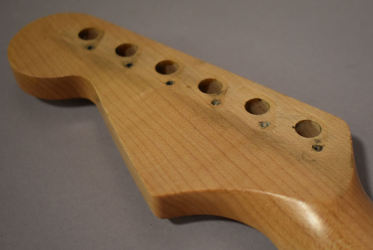 The maple Stratocaster neck's headstock is angled right, showing six tuner holes with screw marks and light wear on the natural wood finish.