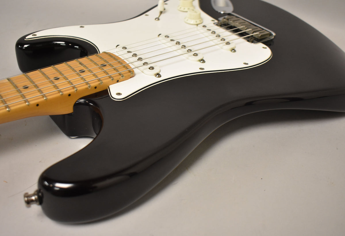 Angled view of a glossy black Stratocaster body with a white pickguard, three single-coil pickups, and a maple neck.