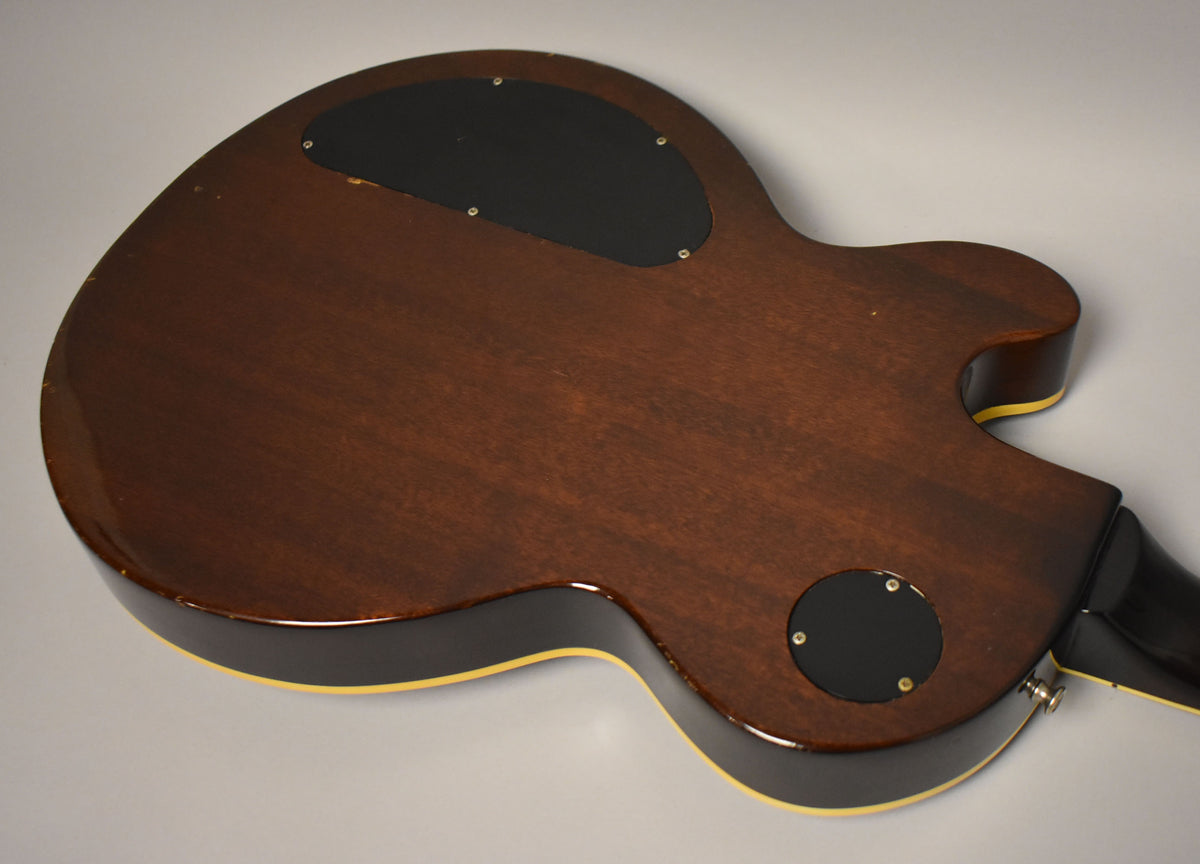 The back of a dark brown guitar body, showing two black control cavity covers, a strap button, and some finish wear.