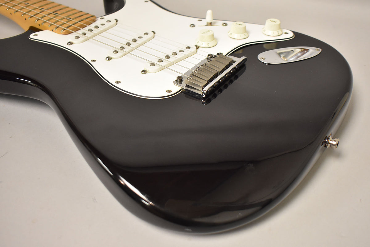 Close-up of the black Stratocaster body, showing the white pickguard, three single-coil pickups, chrome bridge, and output jack.