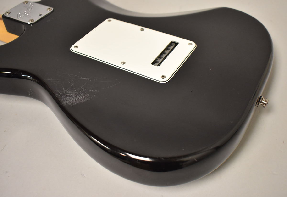 The black Stratocaster's back shows a white tremolo cover, output jack, and heavy surface scratches on the upper bout.