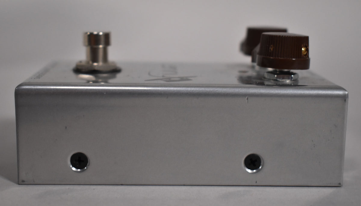 The silver pedal is viewed from the right side, showing two dark brown knobs, a silver footswitch, and two black screws.