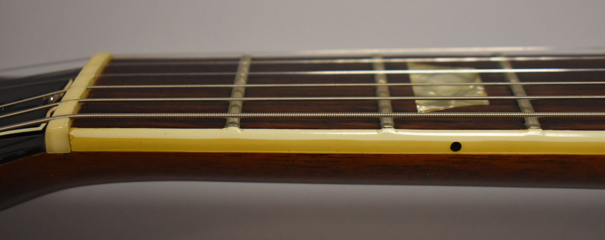 Close-up of the dark walnut fretboard and neck, showing the cream binding, nut, frets, and trapezoid inlay at the 3rd fret.