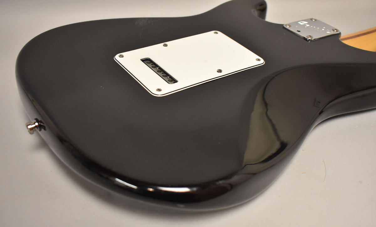 The black Stratocaster's glossy back shows a white tremolo cover, output jack, and light surface wear on the upper horn.