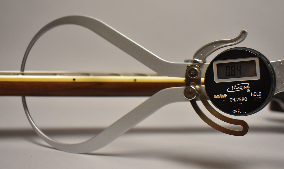 A digital caliper measures the dark walnut neck, showing a reading of 0.84, with cream binding and fret markers visible.