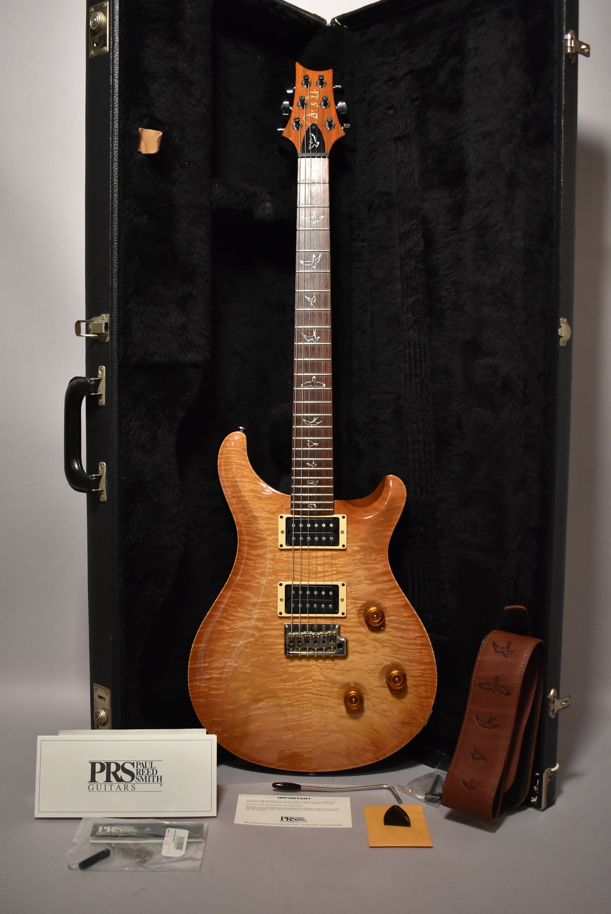 Full view of a sunburst electric guitar with a flamed maple top, gold hardware, and bird inlays, resting in its open black hard case.