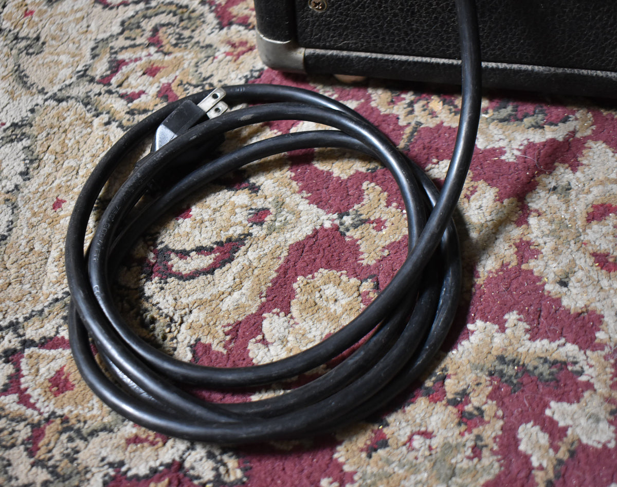 The black power cable, coiled on a patterned rug, extends from the lower right corner of the black amplifier cabinet.
