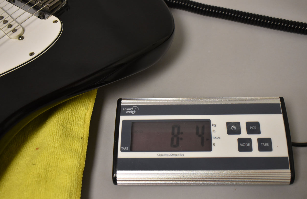 The black guitar body rests on a yellow cloth, with a digital scale displaying "8.4" in the foreground.